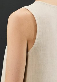 Top sin mangas de color beige claro con tela texturizada, escote redondo y bordes sin terminar, destacando un diseño minimalista.