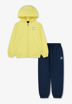 Gul hættejakke med lynlås og forlommer samt navy blå elastiske bukser med talje, begge med små Converse All Star-logoer.