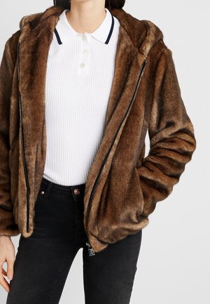 Person trägt eine braune Kunstpelz-Kapuzenjacke über einem weißen, gerippten Poloshirt und schwarzen Jeans, mit einer Hand in der Jackentasche.