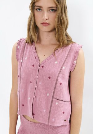 Junge Frau mit welligem blondem Haar, die eine ärmellose pinke Bluse mit Rüschen, Knöpfen und kleinen bestickten Blumenmustern trägt.