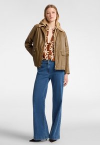 Chaqueta marrón ligera con capucha, combinada con una blusa de estampado y jeans de pierna ancha azules. Presenta cierres de cremallera y botón, y bolsillos de parche.