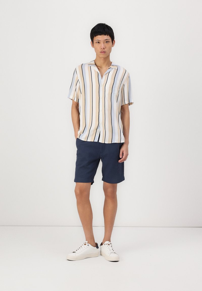 Short-sleeve shirt with vertical beige and navy stripes, paired with navy shorts. The model wears white sneakers, set against a plain background.