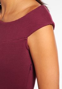 Top a coste, senza spalline, in tessuto bordeaux scuro con una texture sottile; presenta una piccola zip laterale per la chiusura.