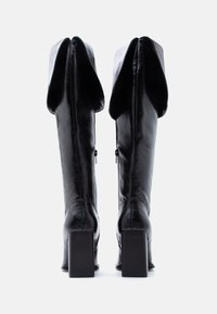 Paire de bottes hautes en cuir noir avec revers pliés et talons blocs, vues de dos sur fond blanc.