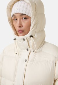 Vrouw in een crèmekleurige pufferjack met de capuchon op en een witte gebreide beanie met een rond logo.