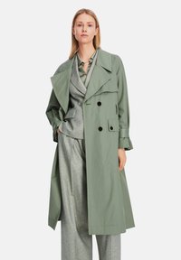 Olive green trench coat with a double-breasted design, large lapels, and button accents, worn over a gray suit with textured fabric.