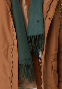 Bufanda de lana verde con bordes desflecados, que presenta un pequeño logo de un árbol bordado. Lleva puesta sobre una chaqueta marrón de cremallera con un tejido texturizado.