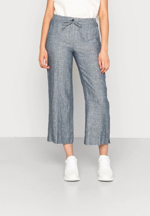 Femme portant un pantalon large gris raccourci avec cordon de serrage et fermeture par bouton, associé à des baskets blanches et un haut crème.