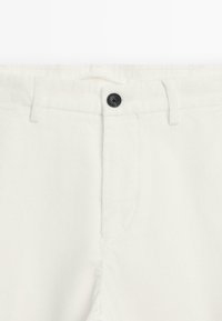 Pantalon en velours côtelé blanc avec un bouton noir à la taille, présentant une coupe droite et des rainures verticales subtiles dans le tissu.