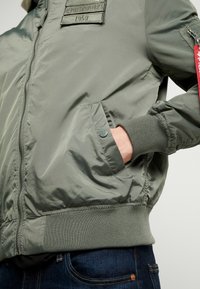 Vihreä pommikenttä takki, joka on valmistettu sileästä kankaasta. Takissa on ribatut hihansuut, vetoketjulliset taskut ja rintamerkki, jossa on teksti "Alpha Industries 1959".