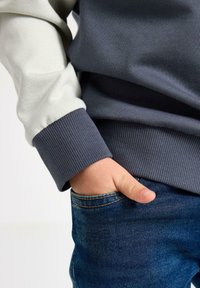 Felpa color block grigia e beige chiaro, polsini a coste, con una texture liscia. Mano appoggiata nella tasca di denim. Design semplice.