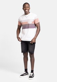 Kortermet, hvit t-skjorte med pastellfargede horisontale striper, lomme-detalj, kombinert med svarte denimshorts og svarte sneakers.