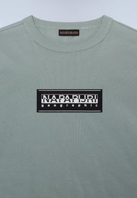 Šviesiai žalias medvilninis marškinėlis su apvaliu kaklu, kuriame yra ryški stačiakampė juodai balta logotipo etiketė su užrašu "NAPAPIJRI geographic."