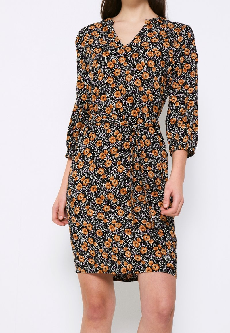 Femme portant une robe noire mi-longue avec un motif floral orange, col en V, manches trois-quarts et une ceinture nouée à la taille.