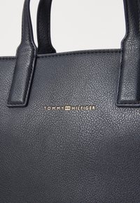 Sac à main en cuir bleu marine avec surface texturée, affichant la marque dorée "TOMMY HILFIGER", poignées supérieures doubles et piqûres ton sur ton.