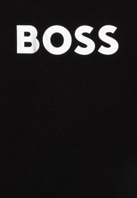 T-shirt noir avec un grand texte blanc en gras qui indique "BOSS" centré sur le devant. Le matériau semble doux et lisse.