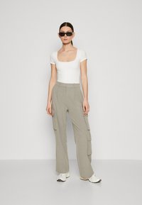 Hvit kortermet tettsittende topp, grå vide cargo-bukser med sidelommer, brukt med svarte solbriller og hvite sneakers på en ensfarget bakgrunn.