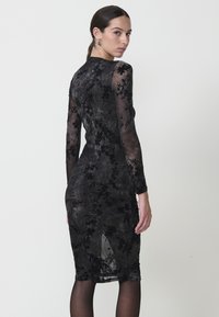 Rochie din dantelă florală neagră, cu mâneci lungi și o siluetă ajustată. Overlay transparent cu un finisaj ușor texturat și tăietură până la genunchi.