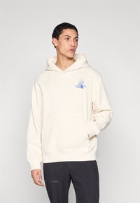 Kremasti pulover s kapuljačom s prednjim džepom, podesivom kapuljačom s vezicama i plavim logotipom na prsima, izrađen od mekanog tekstilnog materijala.