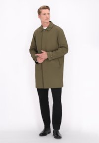 Oliven grøn trenchcoat med skårede krave, knaplukning og sidelommer. Bæret over sorte bukser og ankelstøvler. Glat tekstur.