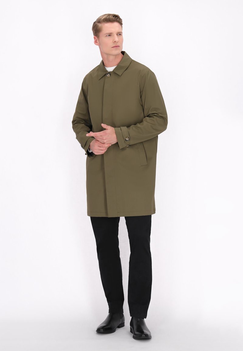 Oliven grøn trenchcoat med skårede krave, knaplukning og sidelommer. Bæret over sorte bukser og ankelstøvler. Glat tekstur.