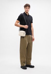 Čierne polo tričko, voľné khaki nohavice, čierne čižmy a malá béžová taška na rameno so zipsom. Jednoduchý, funkčný dizajn.