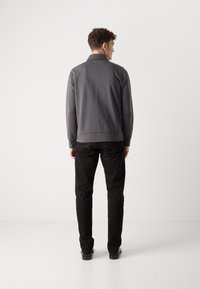 Giacca grigia con una texture liscia, polsini a costine e colletto, abbinata a pantaloni e scarpe nere, vista da dietro.