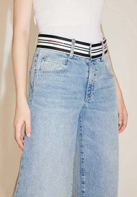 Jeans de mezclilla de pierna ancha en un tono azul claro con una cintura alta. Combinados con una cinturilla elástica de rayas blancas y negras que lleva el logo de la marca.