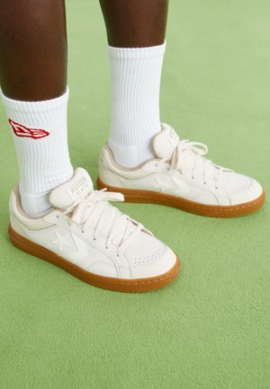 Cremefarbene Ledersneaker mit beiger Gummisohle und dezentes Sternmuster, kombiniert mit weißen gerippten Socken mit roten Logoakzenten.