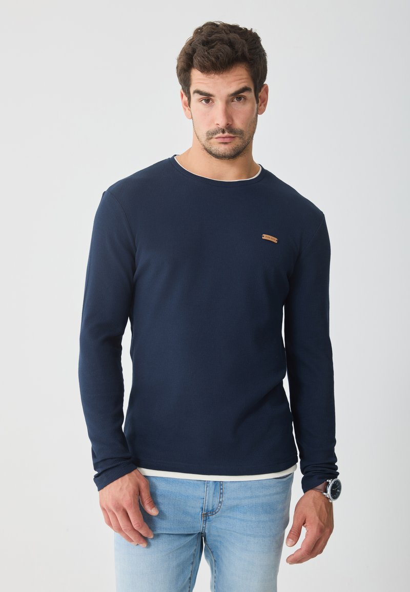 Mornarska majica dolgih rokavov z ribasto teksturo, okroglo ovratnikom, belim poudarkom na ovratniku in majhno rjavo logotipno podrobnostjo na prsih.