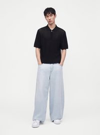 Jeune homme debout, mains dans les poches, portant un polo noir à manches courtes, un jean large bleu clair et des baskets blanches sur fond blanc.