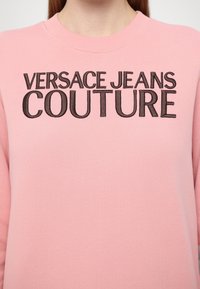Rozā džemperis ar reljefām melnām "VERSACE JEANS COUTURE" burtiem uz krūtīm. Gluda auduma ar apaļu izgriezumu un klasisku piegriezumu.