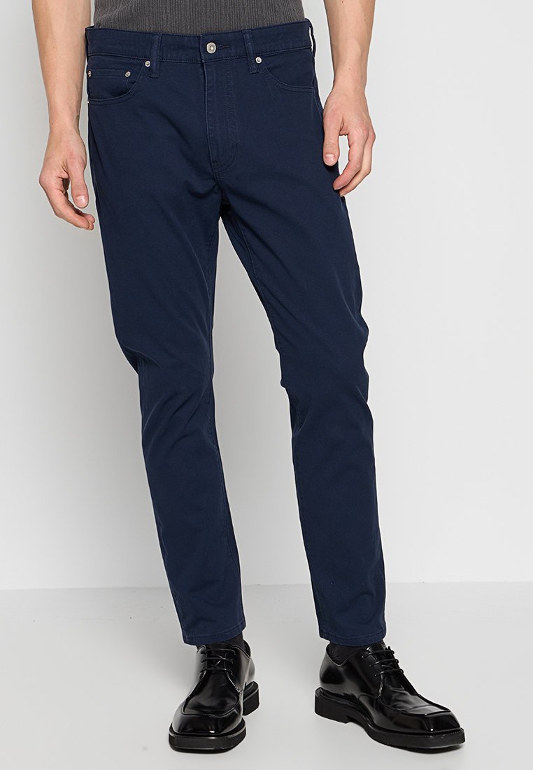 Man in een marineblauwe slim-fit broek met voorzakken en zwarte gepolijste nette schoenen tegen een effen lichte achtergrond.