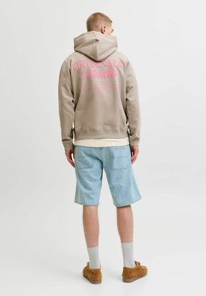 Man draagt een beige hoodie met roze tekst, lichtblauwe denimshorts, witte sokken en bruine schoenen, staand met de rug naar de camera tegen een witte achtergrond.