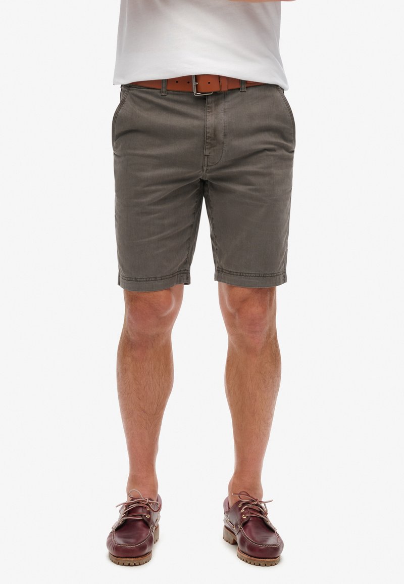 Pantalones cortos de algodón grises con un corte clásico y bolsillos frontales, combinados con un cinturón de cuero marrón. El modelo lleva zapatos de barco marrones.