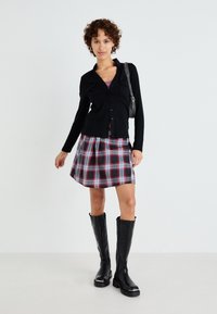 Cárdigan negro de manga larga con botones, combinado con una falda plisada de cuadros en rojo, negro y blanco, y botas negras hasta la rodilla.