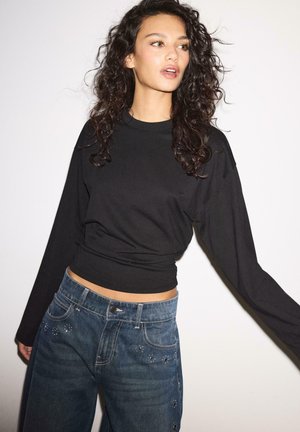 Ženska s kodrastimi temnimi lasmi, oblečena v črno majico z dolgimi rokavi in modre kavbojke z majhnimi dekorativnimi zakovicami, stoji pred preprostim bež ozadjem.