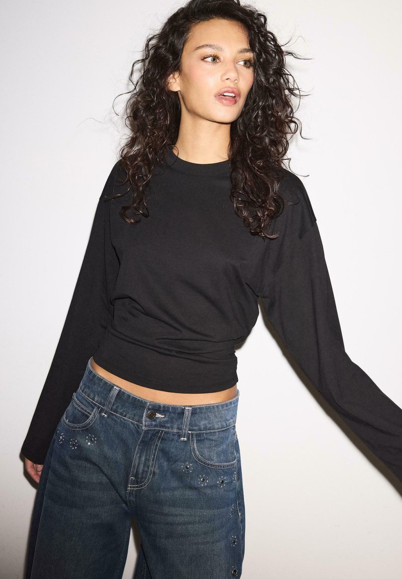 Kvinde med krøllet mørkt hår iført en sort bluse med lange ærmer og blå jeans med små dekorative nitter, stående foran en ensfarvet hvid baggrund.