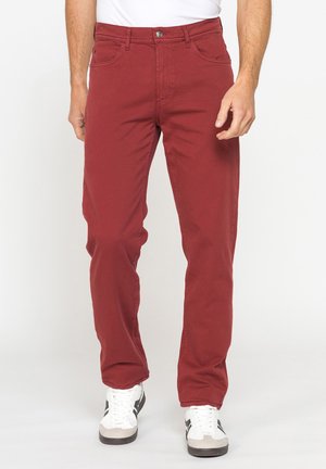 Uomo con pantaloni rossi a gamba dritta e sneaker bianche in piedi davanti a uno sfondo bianco uniforme.