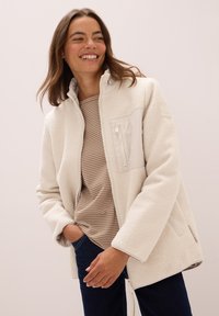 Veste en polaire blanche avec un col haut et une poche poitrine zippée. Chemise rayée beige en dessous, associée à un jean bleu foncé.