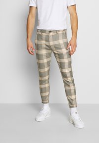 Man som bär slim-fit byxor i grå och orange rutigt mönster, vita strumpor, vita Adidas sneakers och en enkel vit T-shirt mot en ljusgrå bakgrund.