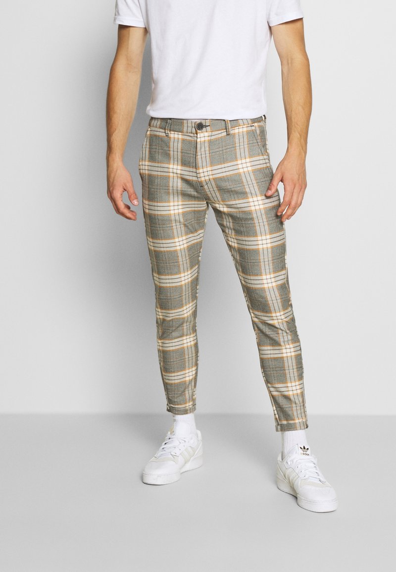 Man som bär slim-fit byxor i grå och orange rutigt mönster, vita strumpor, vita Adidas sneakers och en enkel vit T-shirt mot en ljusgrå bakgrund.