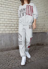 Set di felpa grigia e pantaloni sportivi con testo audace in rosso scuro e nero, dall'aspetto rilassato e dalla consistenza liscia. Sneakers bianche completano il look.