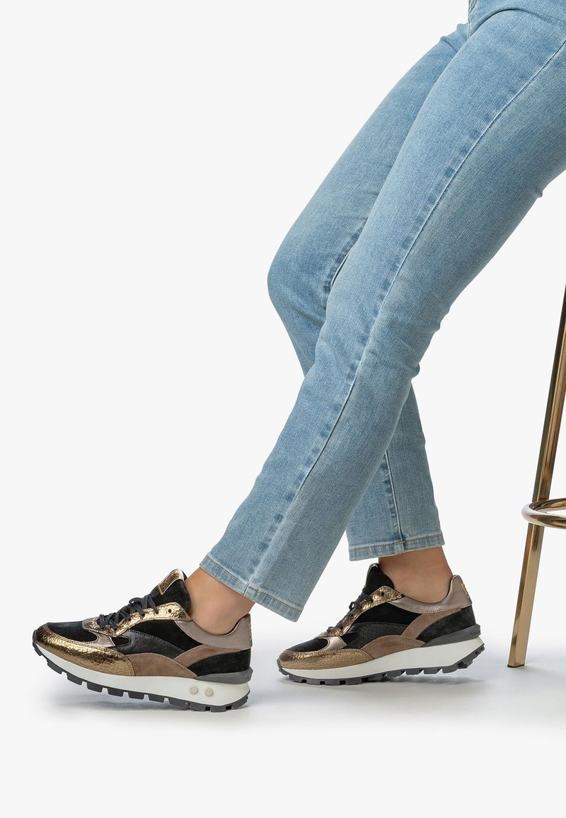 Nogi w jasnoniebieskich dżinsach, ubrane w czarne i metaliczne złote sneakersy, skrzyżowane podczas siedzenia na złotym metalowym stołku na białym tle.