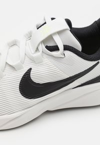 Fehér sportcipő, texturált szövetből, fekete kiegészítőkkel, kiemelkedő fekete swoosh logóval és állítható pánttal a biztonságos illeszkedés érdekében.