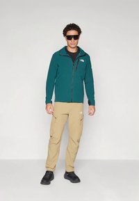 Mann med teal zip-up jacket, beige cargo bukser, sorte sneakers og store sorte solbriller, stående mot en enkel lys bakgrunn.