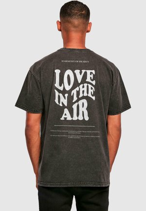 Svart bomull T-skjorte med oversized passform, med hvit tekst som sier "LOVE IN THE AIR" og ekstra trykt tekst under. Korte ermer og rund hals.
