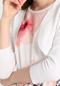 Cardigan blanco y ligero sobre una camiseta con estampado floral en tonos rosas y negros, con un cuello redondo y mangas tres cuartos.