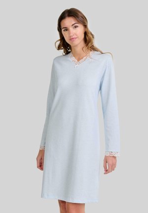 Mujer vestida con un camisón azul claro de mangas largas con encaje en el escote y en los puños, de pie contra un fondo gris plano.