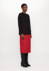 Blusa nera trasparente con dettagli plissettati e maniche lunghe, abbinata a una gonna midi in suede rossa. Stivali in pelle nera fino al ginocchio.